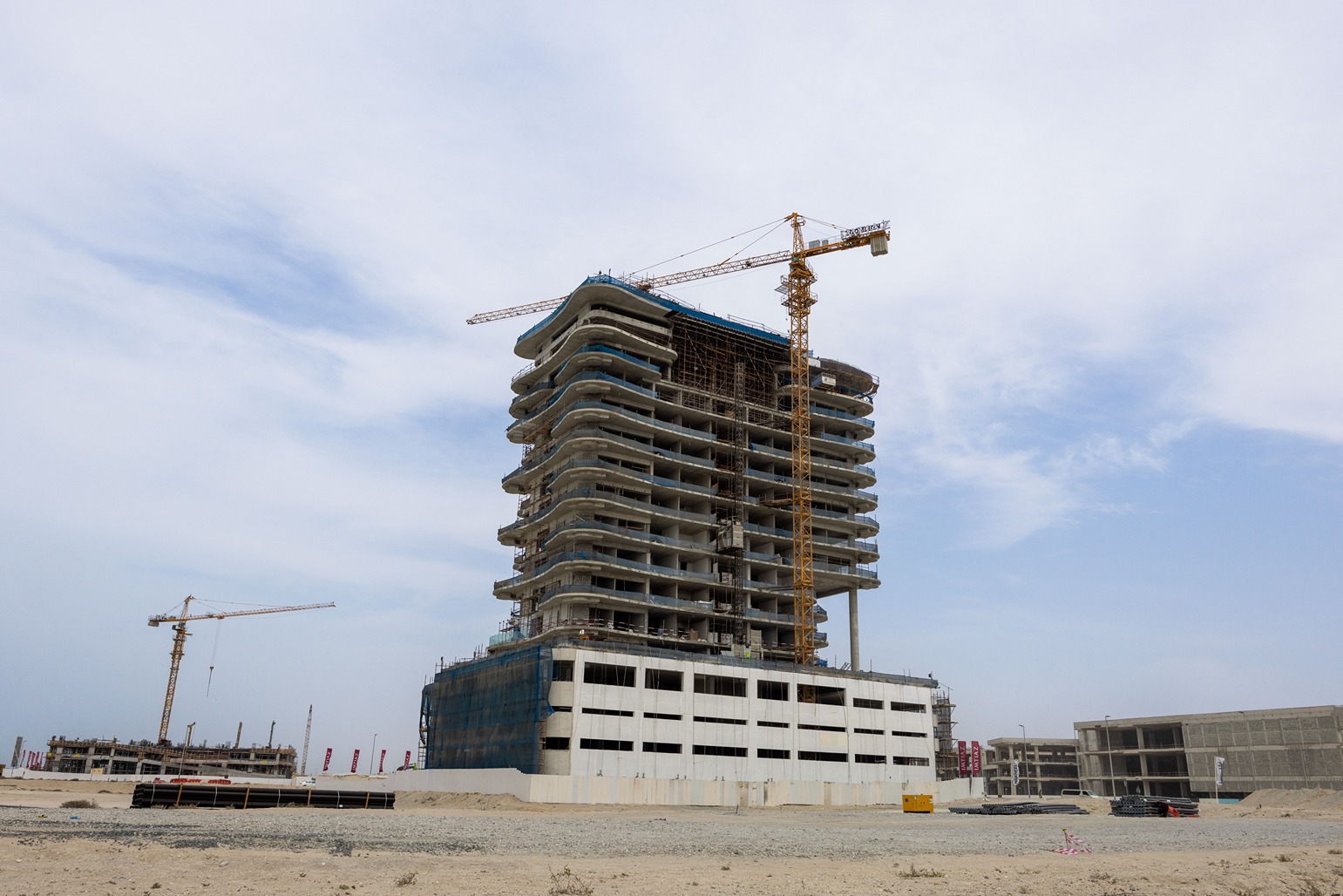 Beach Walk Residence A tall, unfinished building under construction with cranes nearby, set against a clear sky and sandy landscape.